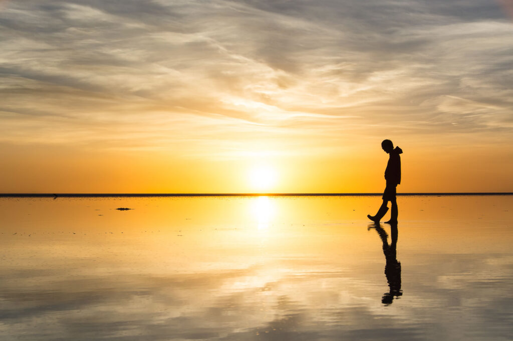 A picture of an orange sunset or sunrise over a  large shallow  pool of water.  A figure is shown in silhouette walking through the water and looking at their reflection.  The person wears a hood thrown back and wading boots.