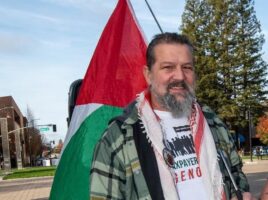 Tarik Kanaana. A Palestinian-American stands holding the Palestinian flag. He has kind eye, a mustache and beard, and trimmed black hair. He wears a plaid flannel shirt over a t-shirt that reads, "Taxpayers Against Genocide."