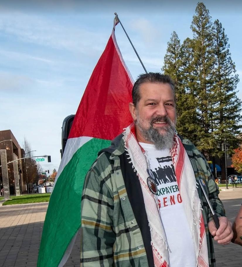 Tarik Kanaana. A Palestinian-American stands holding the Palestinian flag. He has kind eye, a mustache and beard, and trimmed black hair. He wears a plaid flannel shirt over a t-shirt that reads, "Taxpayers Against Genocide."